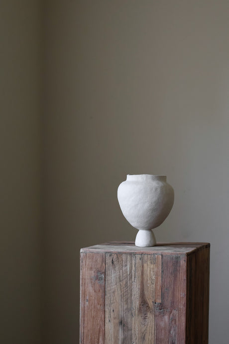 A hand-built, sculptural vase by Hana, crafted from a light-colored clay, leans slightly to the right.  It features a wide, almost spherical body that rests upon a narrow, pedestal-like base. The surface of the vase appears to have a slightly textured, matte finish, indicative of the hand-building process