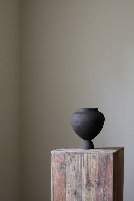 A hand-built, sculptural vase by Hana, crafted from a light-colored clay, leans slightly to the right.  It features a wide, almost spherical body that rests upon a narrow, pedestal-like base. The surface of the vase appears to have a slightly textured, matte finish, indicative of the hand-building process