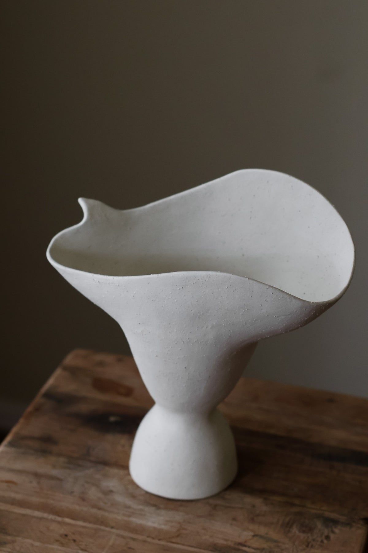 This hand-built ceramic piece exhibits a captivating organic form, reminiscent of a flower blossom gracefully opening. The vase's body begins with a slender base, gradually expanding outward and culminating in a dramatically flared, undulating rim. The rim itself is a work of art, with a gently ruffled edge that evokes the delicate petals of a flower. 
