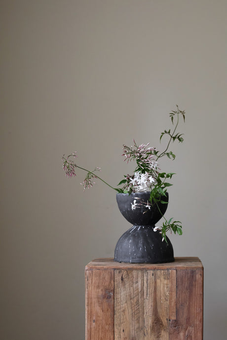Black hand-thrown ceramic hourglass vase by artist Giselle Hicks. The vase features a distinctive hourglass silhouette with a bulbous base and a tapering neck, creating a visually dynamic form. The surface is adorned with a dark, textured glaze, showcasing subtle variations in color and a hand-crafted aesthetic. 